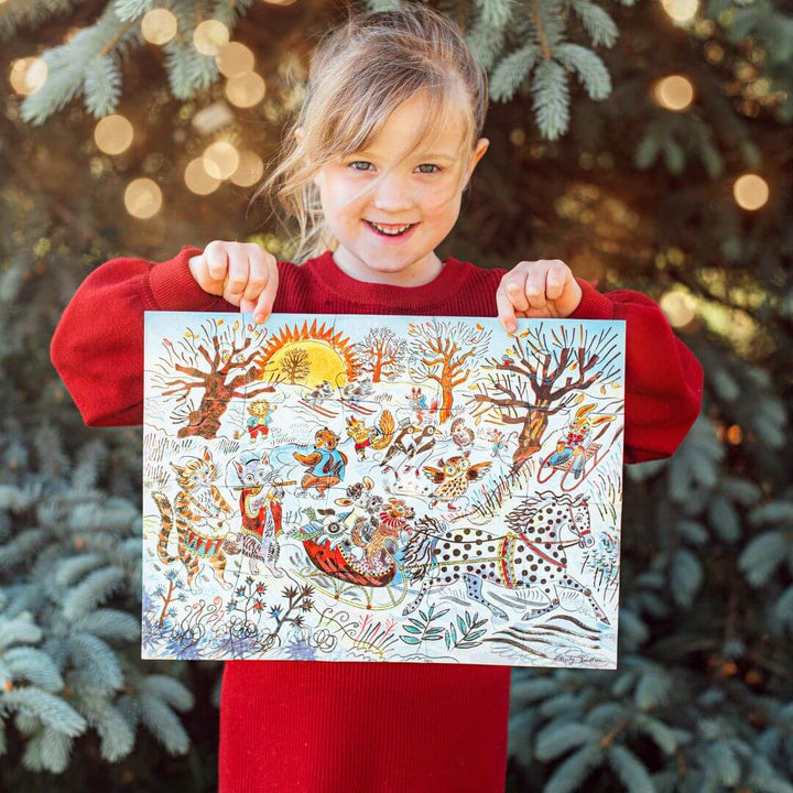 A child holding up a completed eeBoo Winter Sleigh Ride 20-piece puzzle illustrated by Emily Sutton with animals skiing and sledding in a snowy landscape with a lit Christmas tree in the background – Bella Luna Toys.