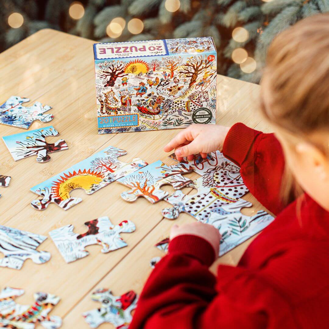 A child sitting at a table assembling eeBoo's Winter Sleigh Ride 20-piece puzzle illustrated by Emily Sutton with animals skiing and sledding in a snowy landscape – Bella Luna Toys.