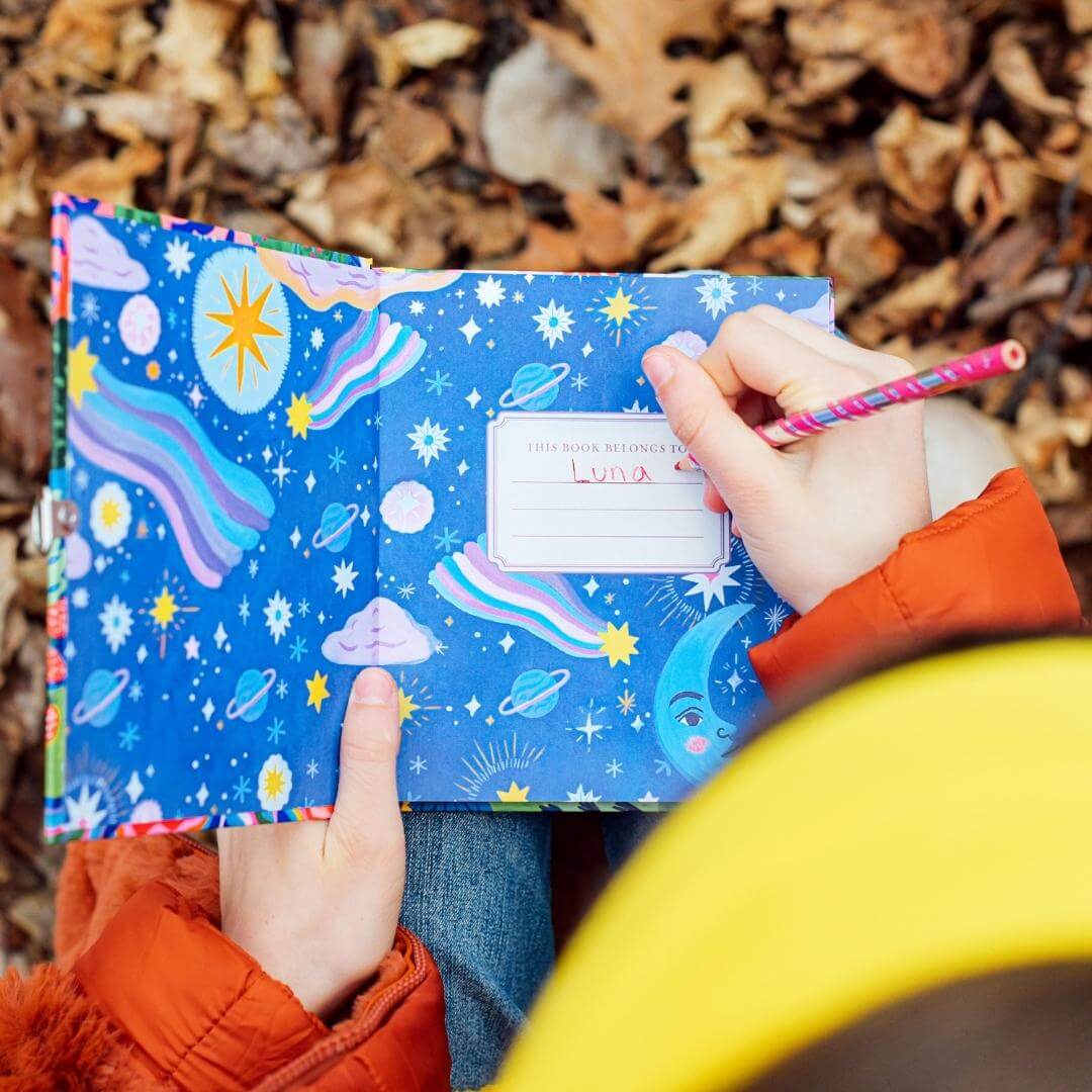 Child writing their name, Luna, in the eeBoo Fawn & Sun Journal with fall leaves in the background.