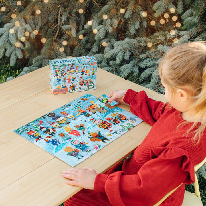 A child sitting at a table assembling the eeBoo Winter Carol 20-piece puzzle illustrated by Yumi Kitagishi with animals and people playing instruments in a winter concert scene – Bella Luna Toys.