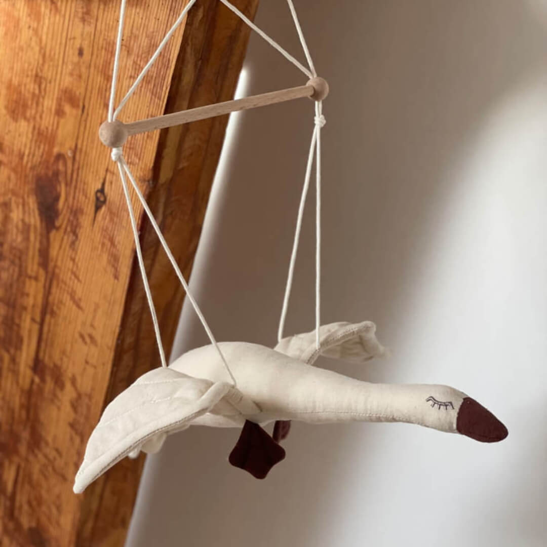 Cotton flying goose mobile with outstretched wings, hanging from a wooden rod with white cords shown with a wooden beam and white wall behind it. 