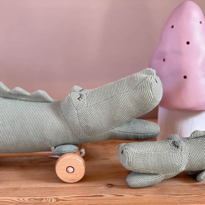 Two plush alligator toys on a wooden surface with a pink background.