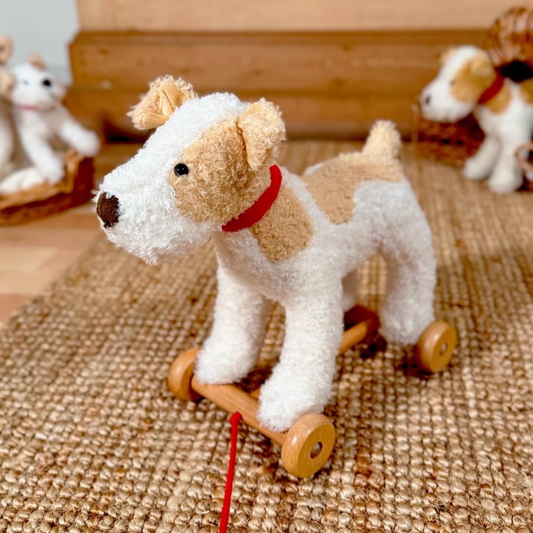 Soft plush Eliot Dog pull-along toy with a red cord and sturdy wooden wheels, designed for toddlers to push or pull during active play shown  on a natural brown rug with other toys in background.