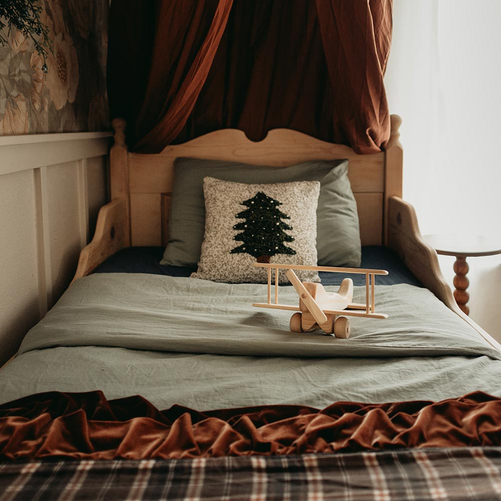 A natural wooden airplane sits on a bed in a soft lit room.