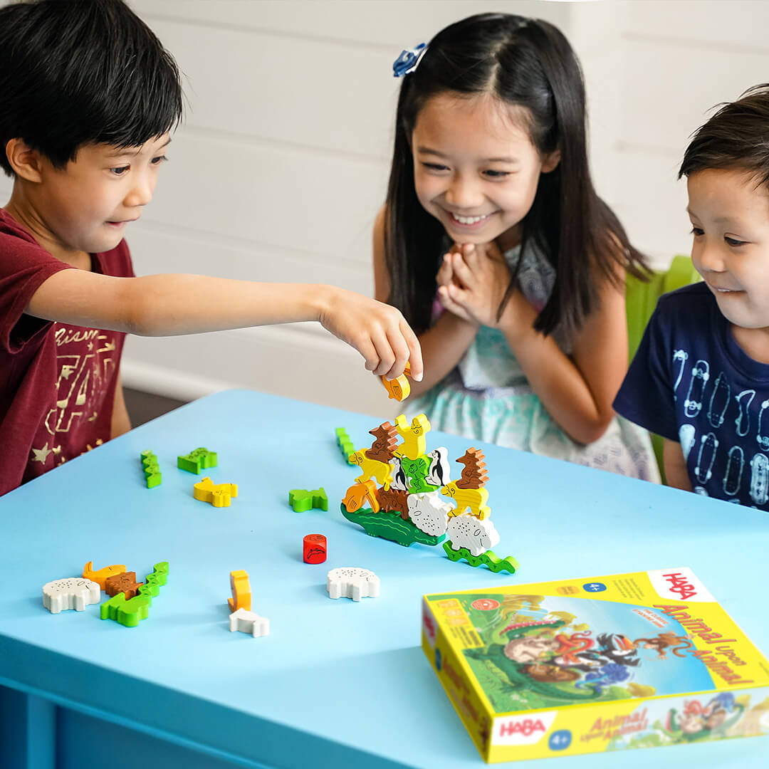 Three children are playing Animal Upon Animal on a blue table, smiling as one places a piece on an animal stack.