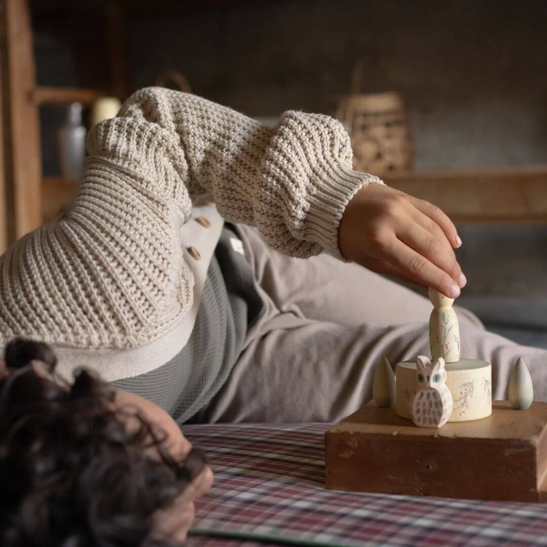 Person wearing knitted sleeves interacting with small wooden toys on a checkered tablecloth. Grapat Hello Winter set