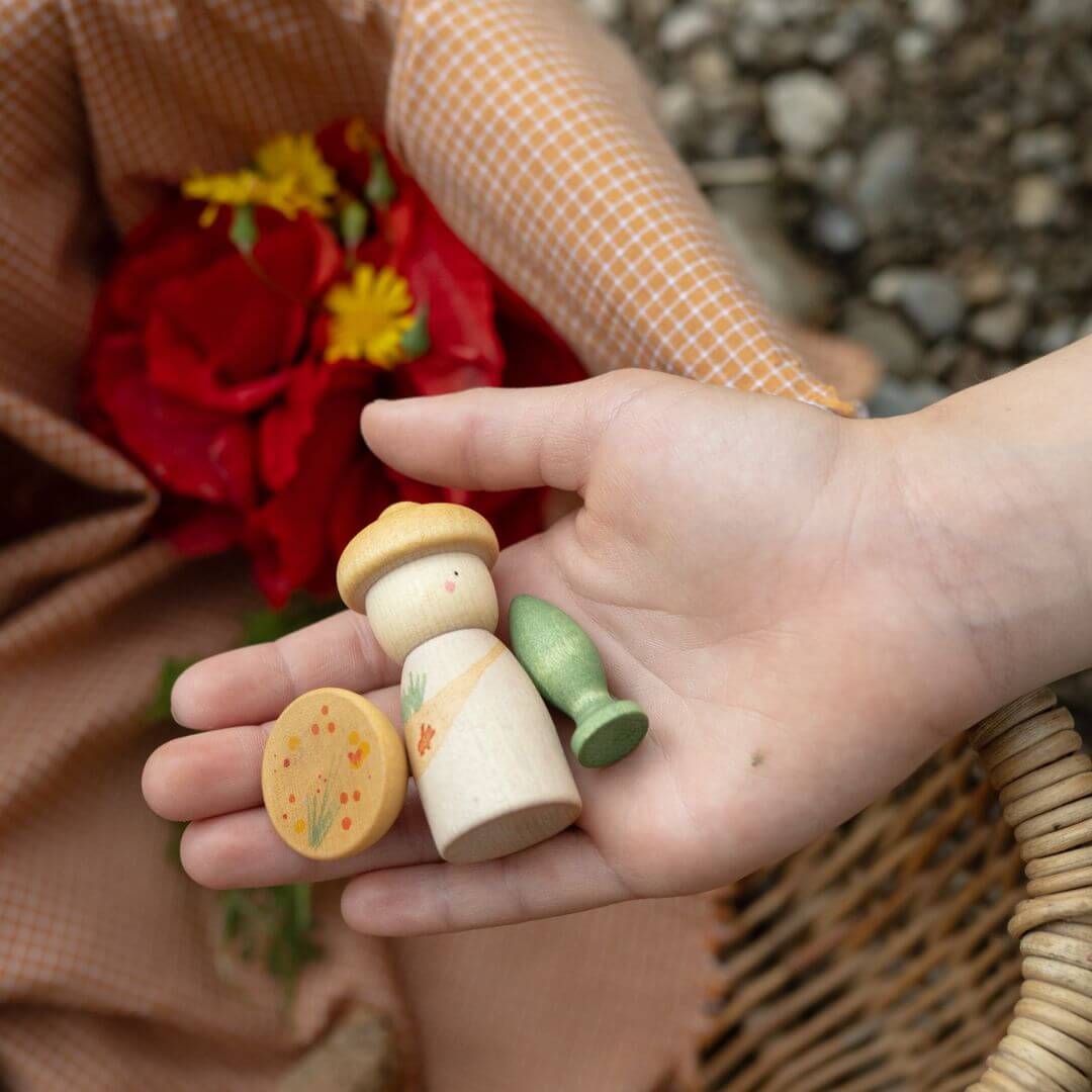 Child holding three figures from one Grapat Lucky Lucky 4 set above a wicker basket and red flowers, highlighting natural textures and play possibilities.
