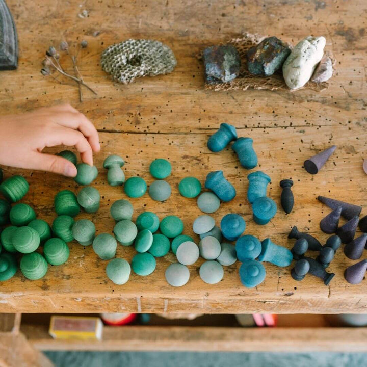A child arranging the Grapat Mandala Mix Cold set with 81 hand-painted wooden mandala pieces in blue, green, and purple tones arranged by color on a wooden table - Bella Luna Toys