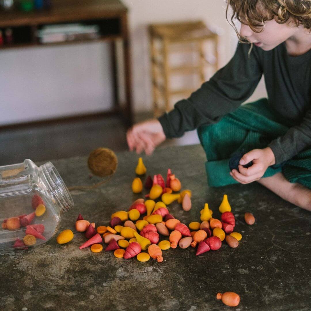 Grapat Mandala Mix Warm set with 81 hand-painted wooden mandala pieces in red, orange, and yellow tones shown spread on a counter with a child playing with them - Bella Luna Toys