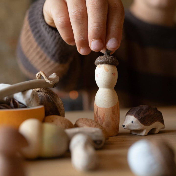 Child placing an acorn cap on a Grapat wooden figure from the Hello Autumn! set, with Ostheimer hedgehog and natural treasures nearby – Bella Luna Toys.