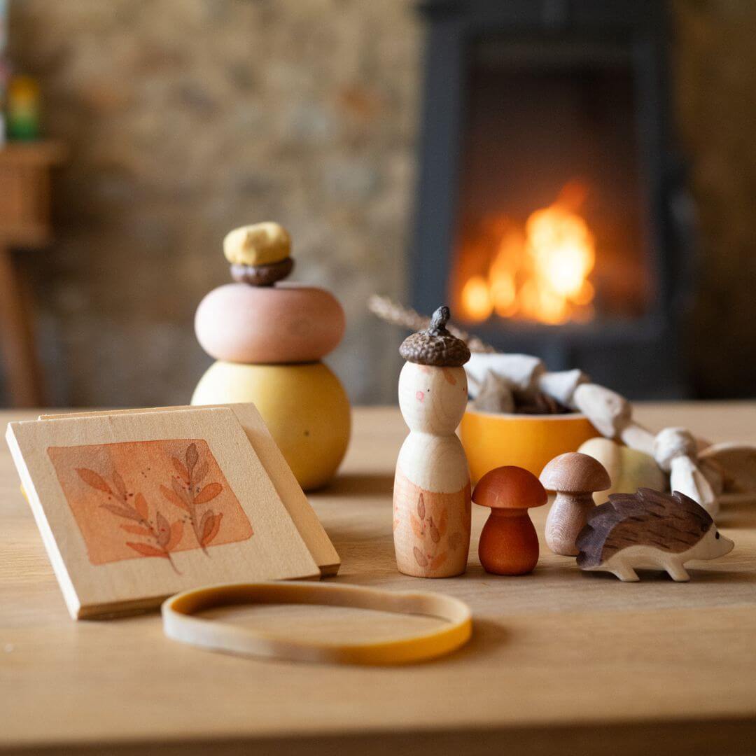 Grapat x Ostheimer Hello Autumn! wooden play set with figure, hedgehog, mushrooms, flower press, and illustrated box displayed on a table with a fire in the background – Bella Luna Toys.