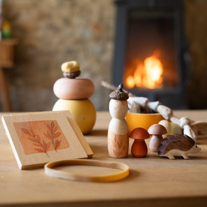 Grapat x Ostheimer Hello Autumn! wooden play set with figure, hedgehog, mushrooms, flower press, and illustrated box displayed on a table with a fire in the background – Bella Luna Toys.
