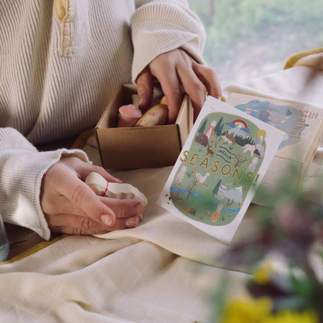 A cozy unboxing moment featuring the Grapat x Ostheimer 'Hello Spring' set. A person wearing a soft cream-colored sweater carefully opens a cardboard box containing wooden seasonal toys. A beautifully illustrated card labeled 'Seasons' with a colorful nature scene is prominently displayed, capturing the essence of seasonal play and storytelling. The warm lighting and natural setting enhance the sense of wonder and connection to nature.
