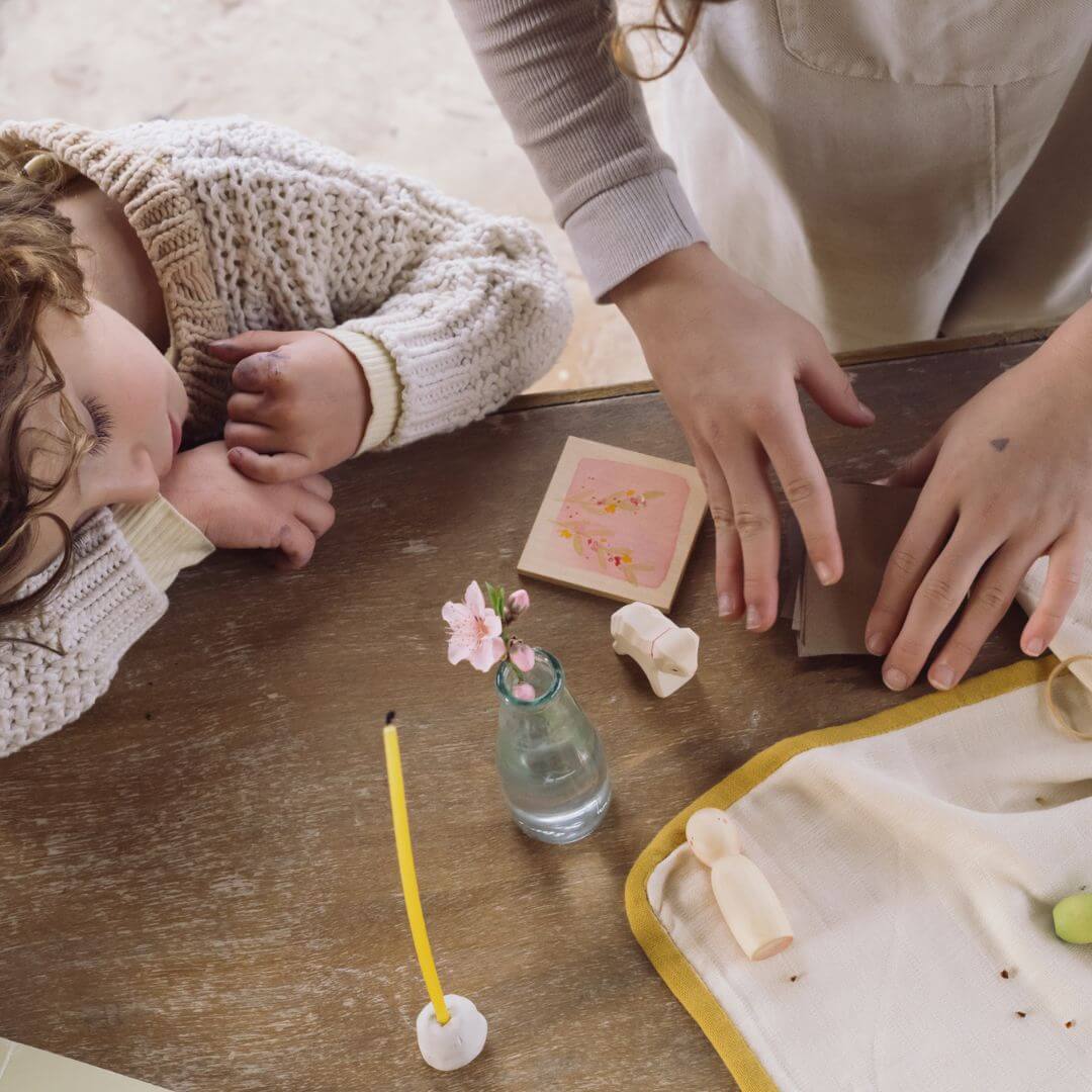 A serene spring-inspired table setting featuring the Grapat x Ostheimer 'Hello Spring' set. The display includes a wooden Nin figure, a miniature Ostheimer lamb, a flower press adorned with blossoms, and small Mandala tulip pieces in pink and green. A glass vase with delicate pink flowers, a beeswax candle, and a young child and and an older child are using the flower press.