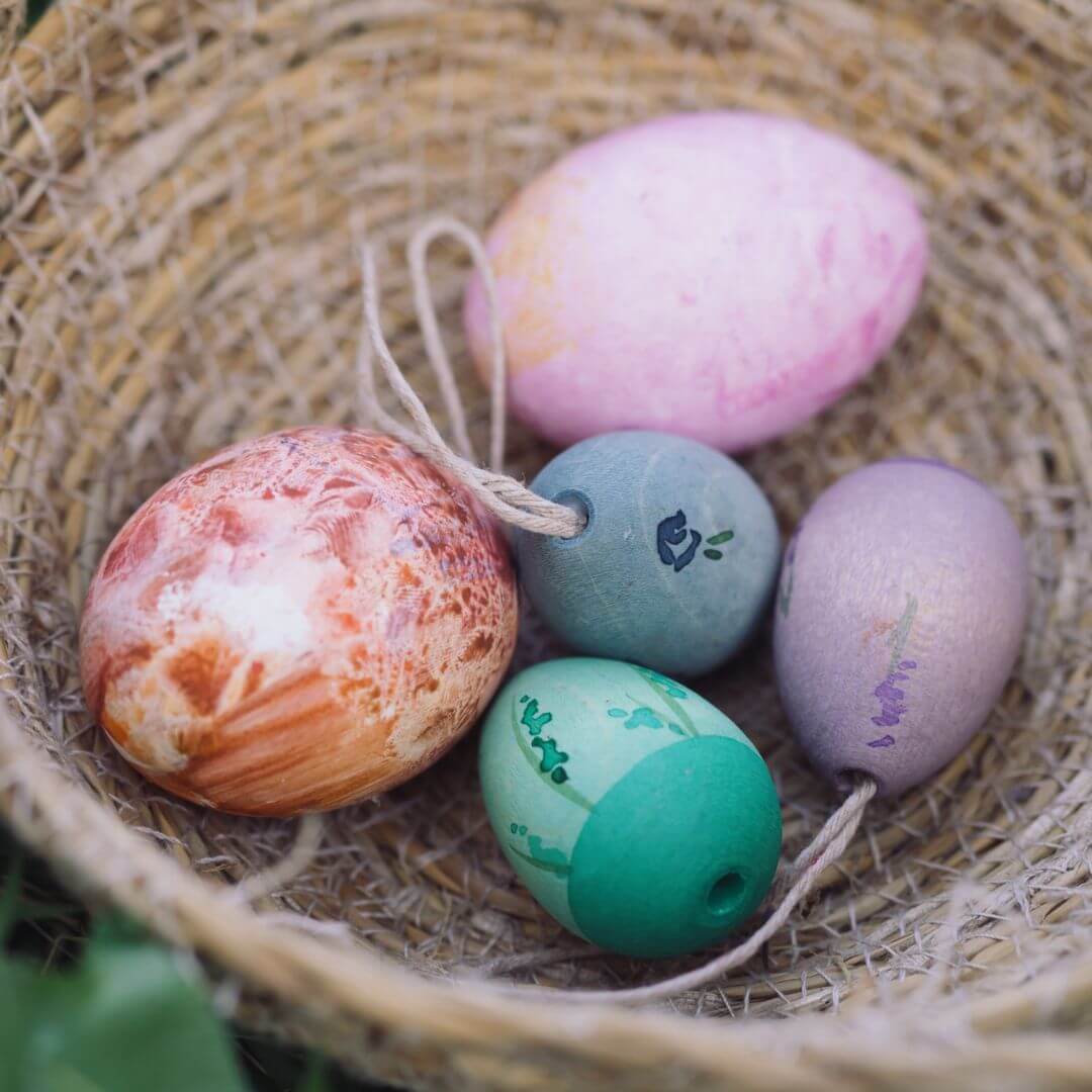 A a basket holds a few handcrafted wooden Easter egg ornaments from Grapat mixed with real hand-painted eggs. The eggs feature cool-toned colors, including blue, green, and purple, with delicate painted details. 