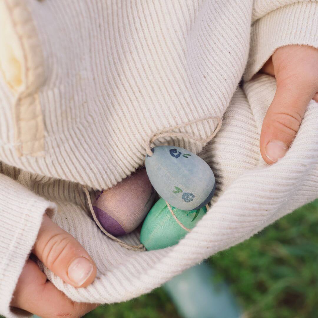 A child gently holds a few handcrafted wooden Easter egg ornaments from Grapat in the fold of their cream-colored sweater. The eggs feature cool-toned colors, including blue, green, and purple, with delicate painted details. The child's small hands and the soft fabric create a cozy and natural atmosphere, highlighting the beauty of these festive decorations.