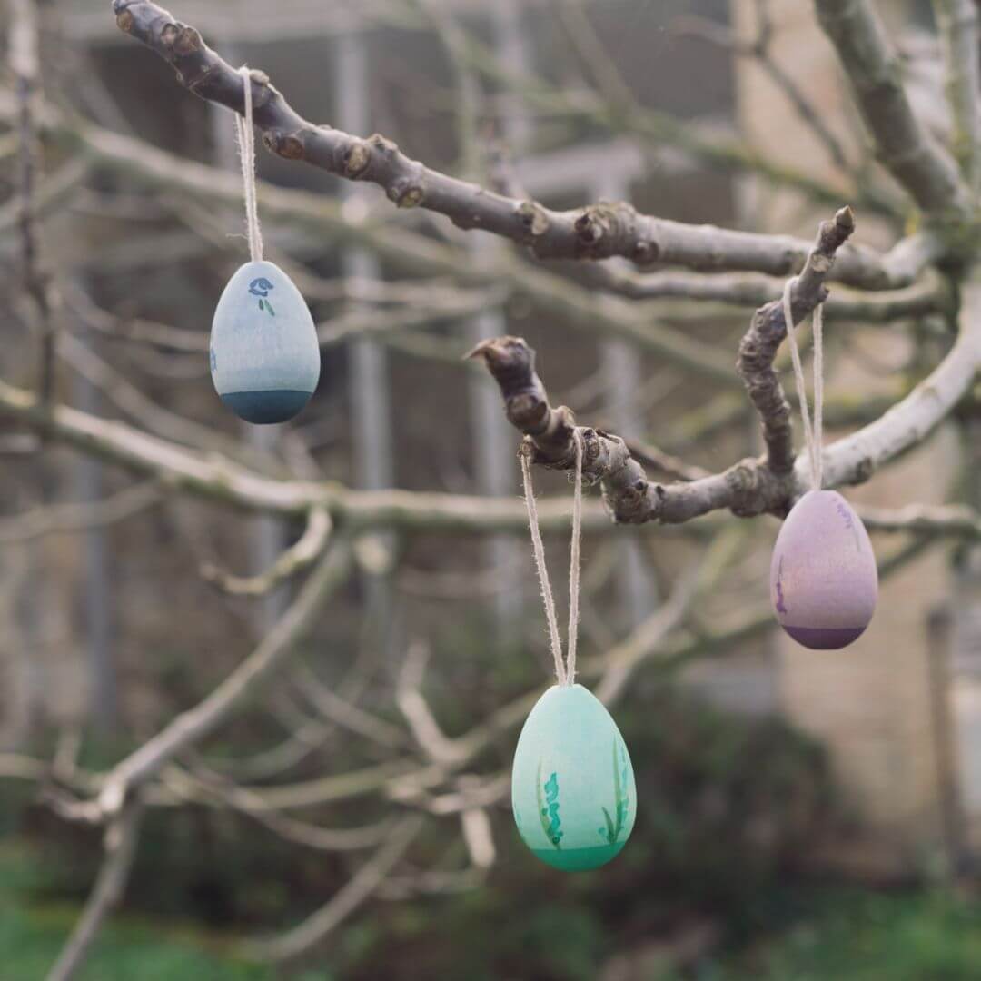 Three handcrafted wooden Easter egg ornaments from Grapat ihang from bare branches of a tree. The eggs feature cool-toned colors, including blue, green, and purple, with delicate painted details. 