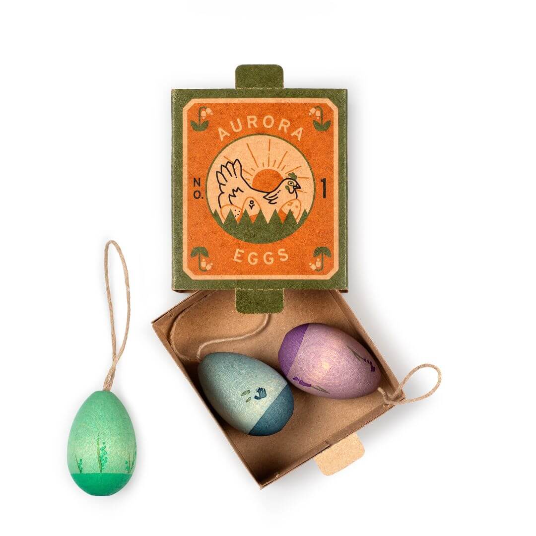 A set of three handcrafted wooden Easter egg ornaments from Grapat, displayed in an open box labeled 'Aurora Eggs No. 1'. The ornaments feature cool-toned colors, including shades of green, blue, and purple, each with a hanging loop for display. The vintage-style packaging showcases an illustration of a rising sun over mountains, adding to the natural and festive Easter theme.