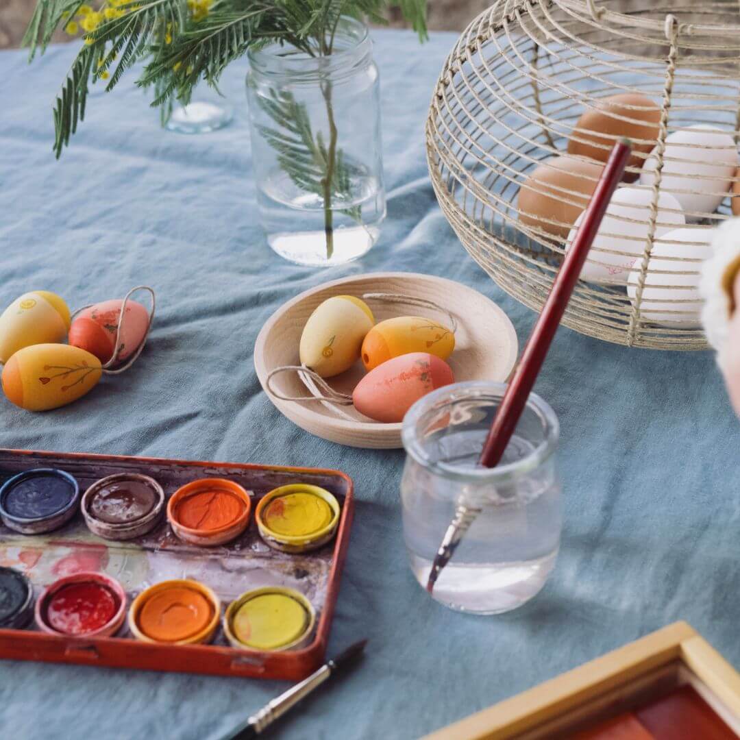 A cozy Easter crafting scene featuring a table covered with a blue cloth. A watercolor paint set, a glass of water with a paintbrush, and a bowl of wooden Easter egg ornaments in warm shades of yellow, orange, and red are arranged on the table. In the background, a wicker basket filled with more eggs and a glass jar with fresh greenery add to the festive and creative atmosphere.