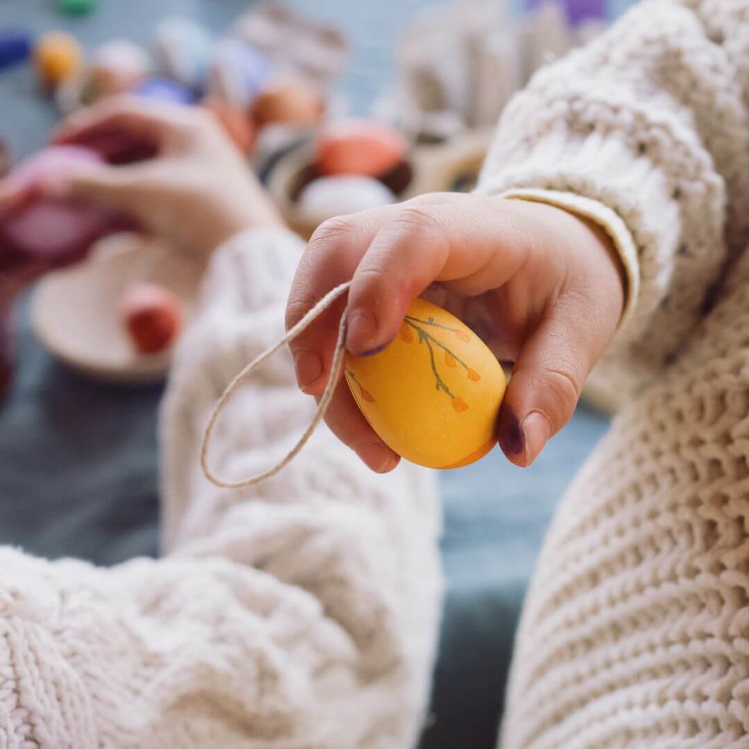 A close-up of a child's hand holding a handcrafted wooden Easter egg ornament from Grapat, painted in a warm yellow shade with delicate decorative details. The egg has a looped string for hanging. In the blurred background, other children and adults are engaged in Easter crafts, creating a cozy and festive atmosphere.