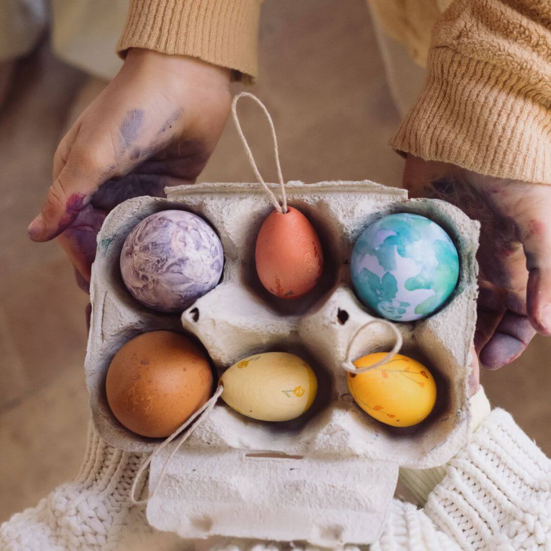 A person wearing a cozy knitted sweater holds a carton of colorful Easter eggs, a mix of handcrafted wooden ornaments and hand-painted real eggs. The eggs display a variety of warm and cool tones, including shades of yellow, orange, blue, and purple. One of the wooden eggs has a looped string for hanging, adding to the festive and artistic Easter presentation.