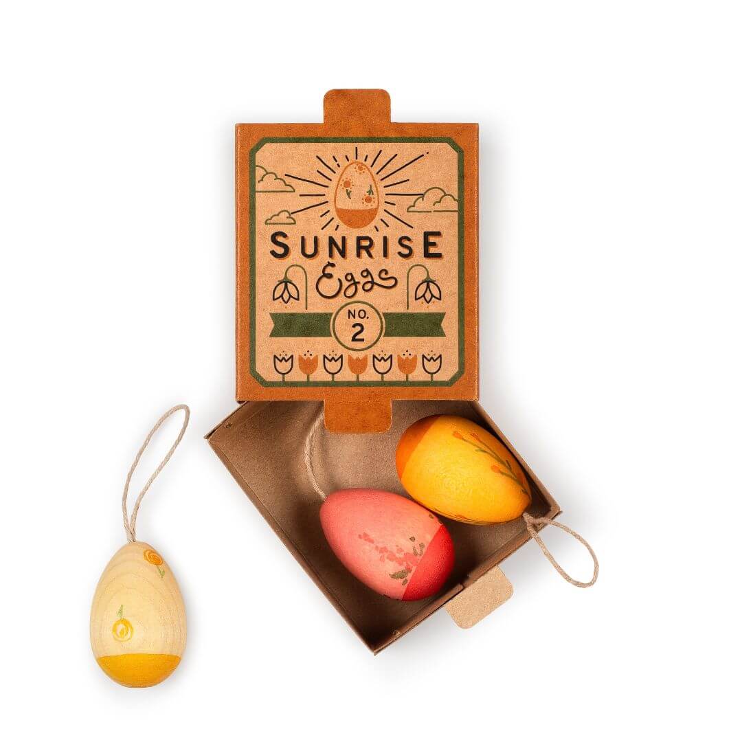 A set of three handcrafted wooden Easter egg ornaments from Grapat, presented in an open box labeled 'Sunrise Eggs No. 2'. The ornaments feature warm, sunrise-inspired colors in shades of yellow, orange, and red, each with a hanging loop for display. The vintage-style packaging includes an illustration of a sun and decorative floral elements, enhancing the festive and natural Easter theme.