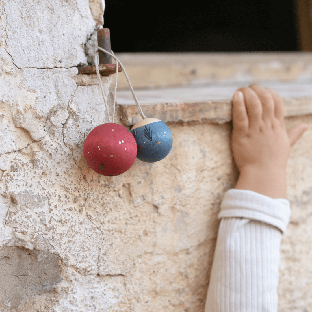 Two Grapat Wish Balls Wooden Ornaments, one red and one blue, hanging from a rustic stone ledge with a child’s hand reaching nearby – Bella Luna Toys.
