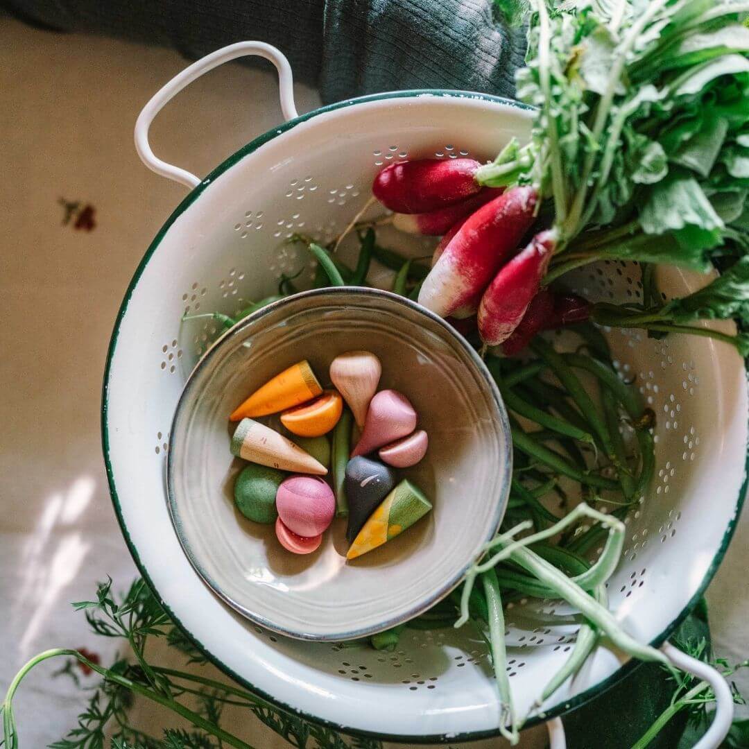 Grapat wooden vegetable play set with 12 hand-painted wooden veggies in assorted shapes and natural colors displayed with real vegetables in a kitchen strainer- Bella Luna Toys