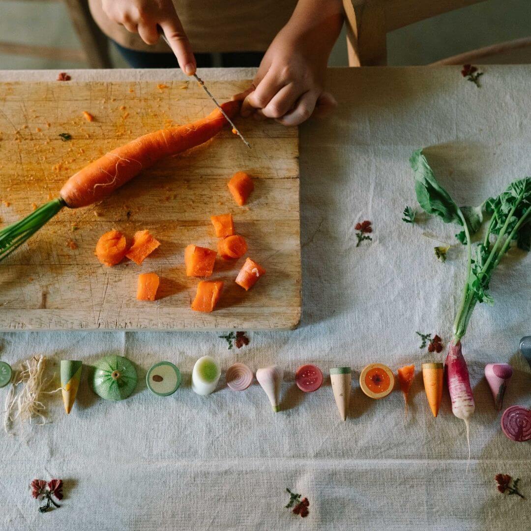 Grapat wooden vegetable play set with 12 hand-painted wooden veggies in assorted shapes and natural colors lined up near a cutting board with someone slicing a carrot - Bella Luna Toys
