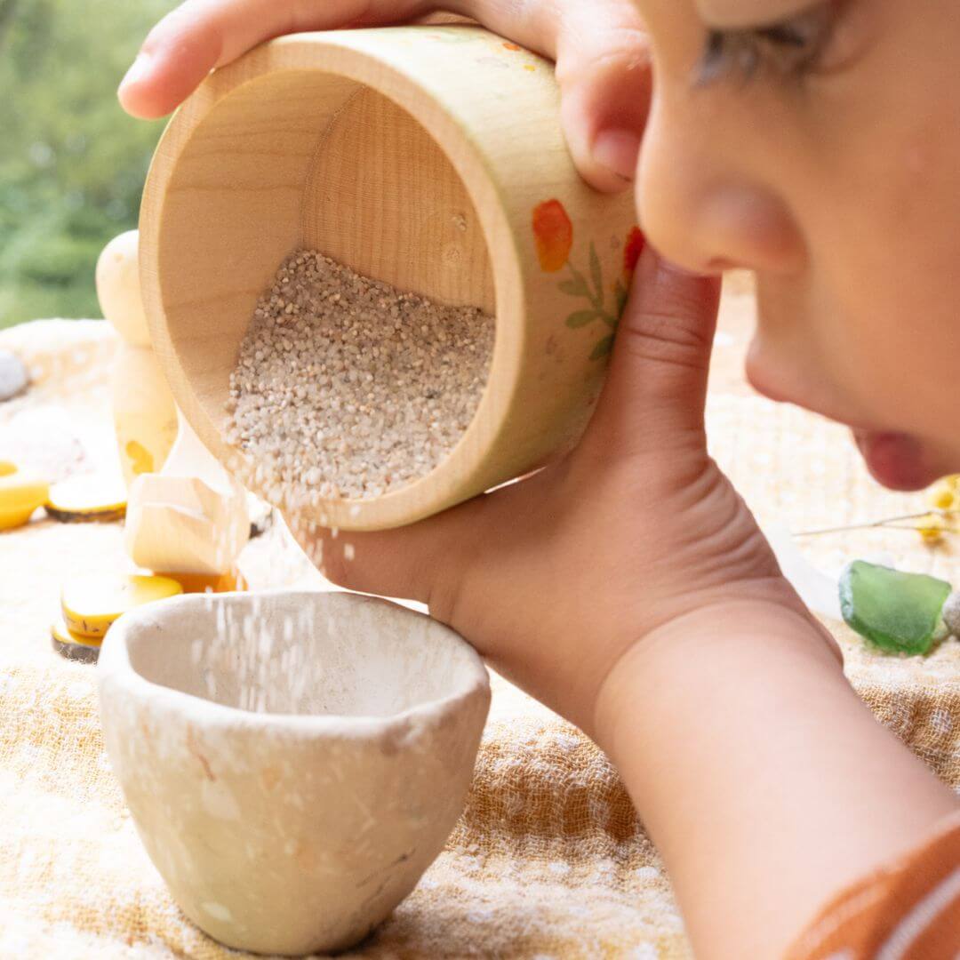 Child playfully pouring sensory materials from the painted wooden bowl included in the Hello Summer Grapat x Ostheimer set, highlighting open-ended play and natural textures.