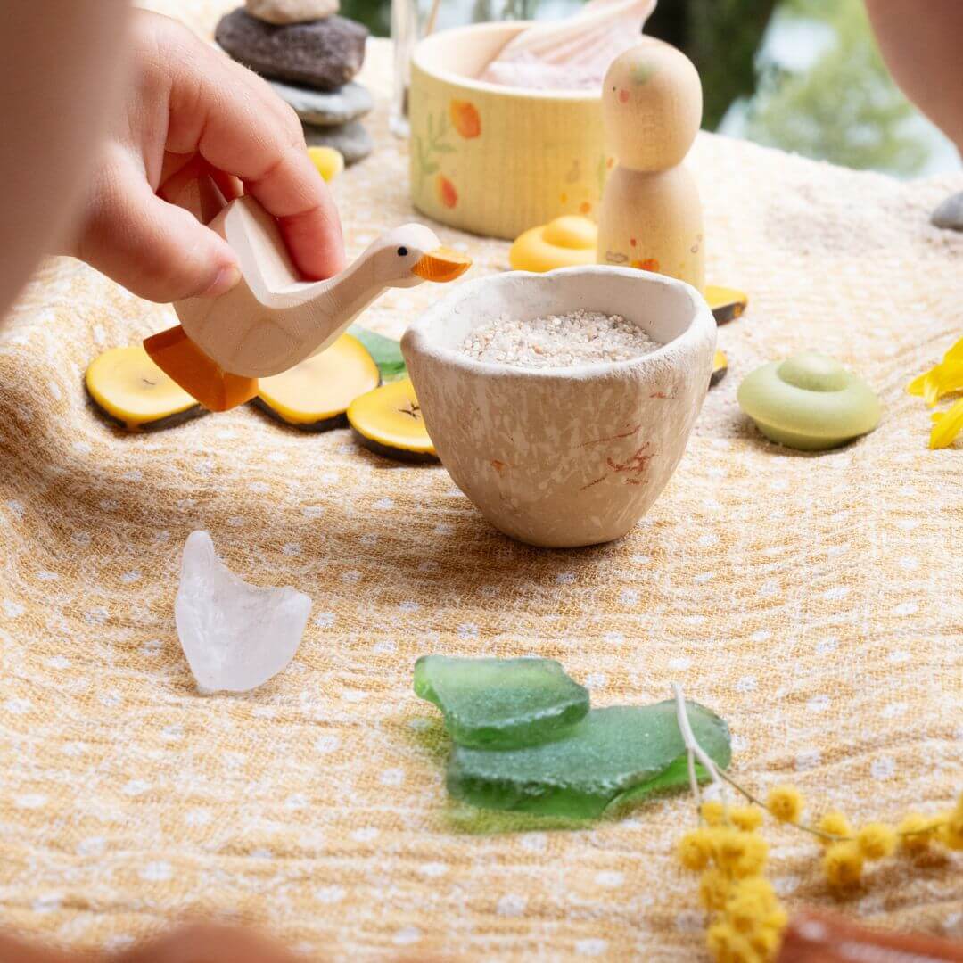 Child’s hand playing with the Ostheimer wooden goose figure from the Hello Summer set, surrounded by natural materials and summer-themed elements on a yellow cloth.
