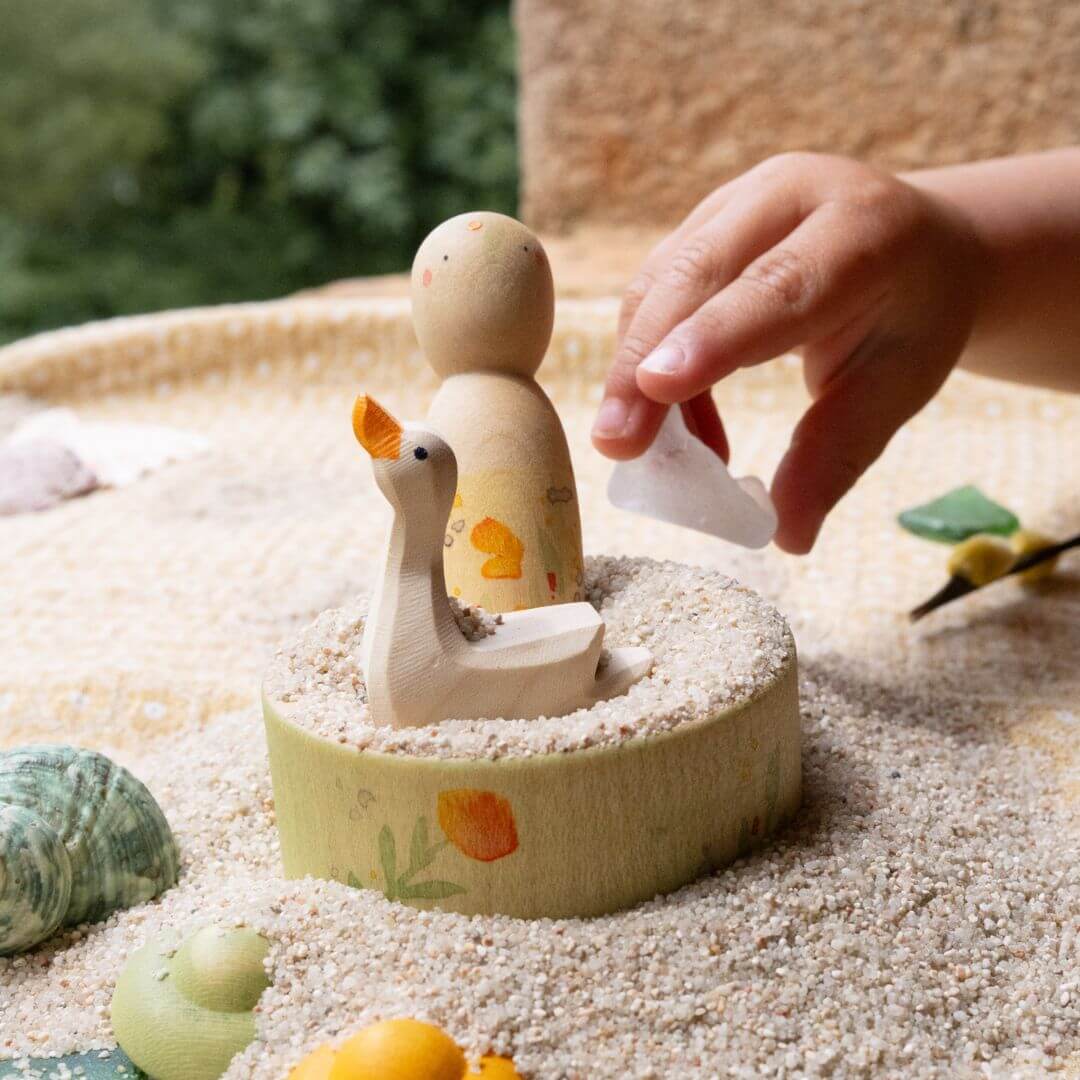 Child’s hand arranging the Grapat Nin and Ostheimer goose figures in a sandbox, using the painted bowl as part of an imaginative summer play scene.