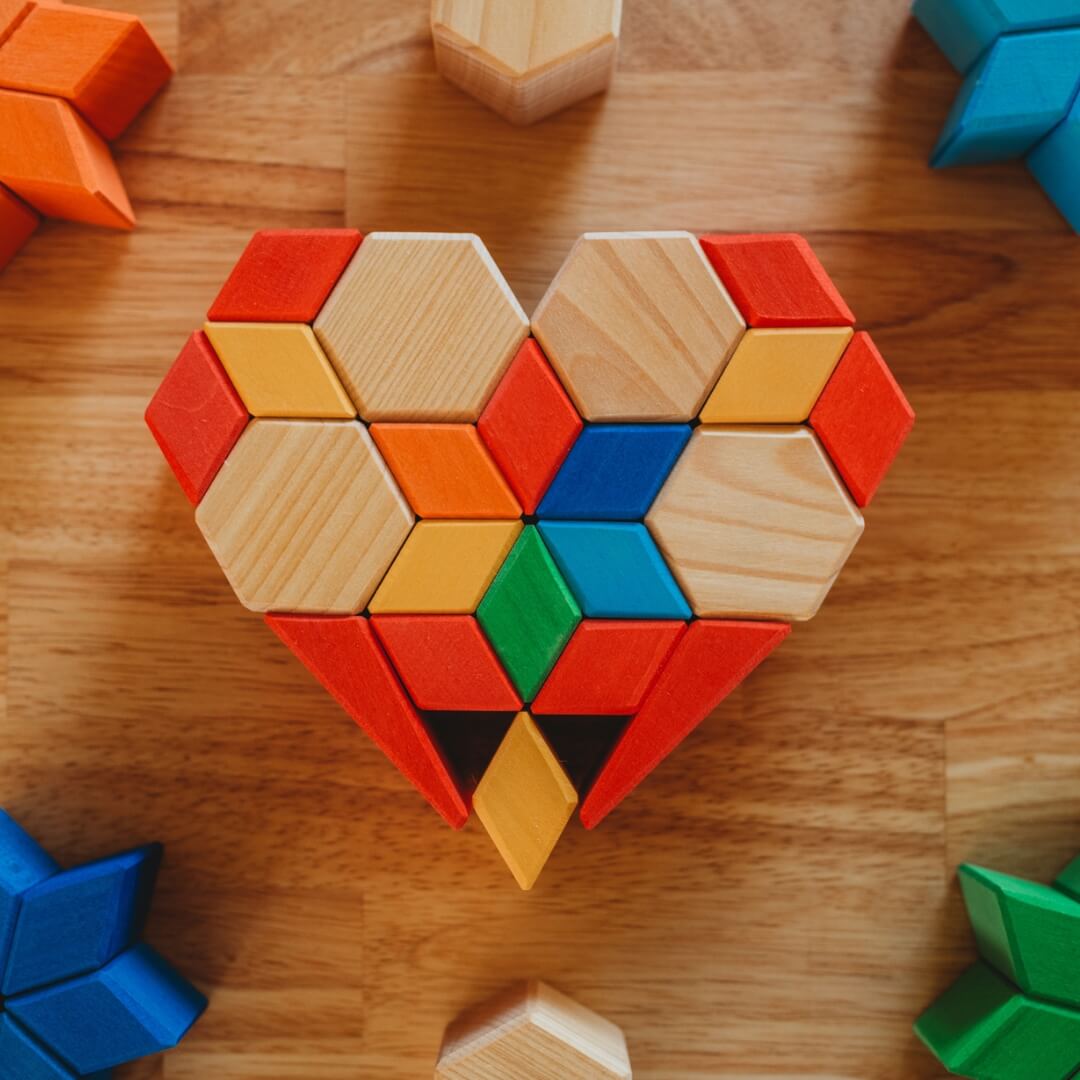 Colorful geometric wooden blocks arranged in a heart shape on a wooden surface. Grimm's Wooden Toys x Bella Luna Toys exclusive.
