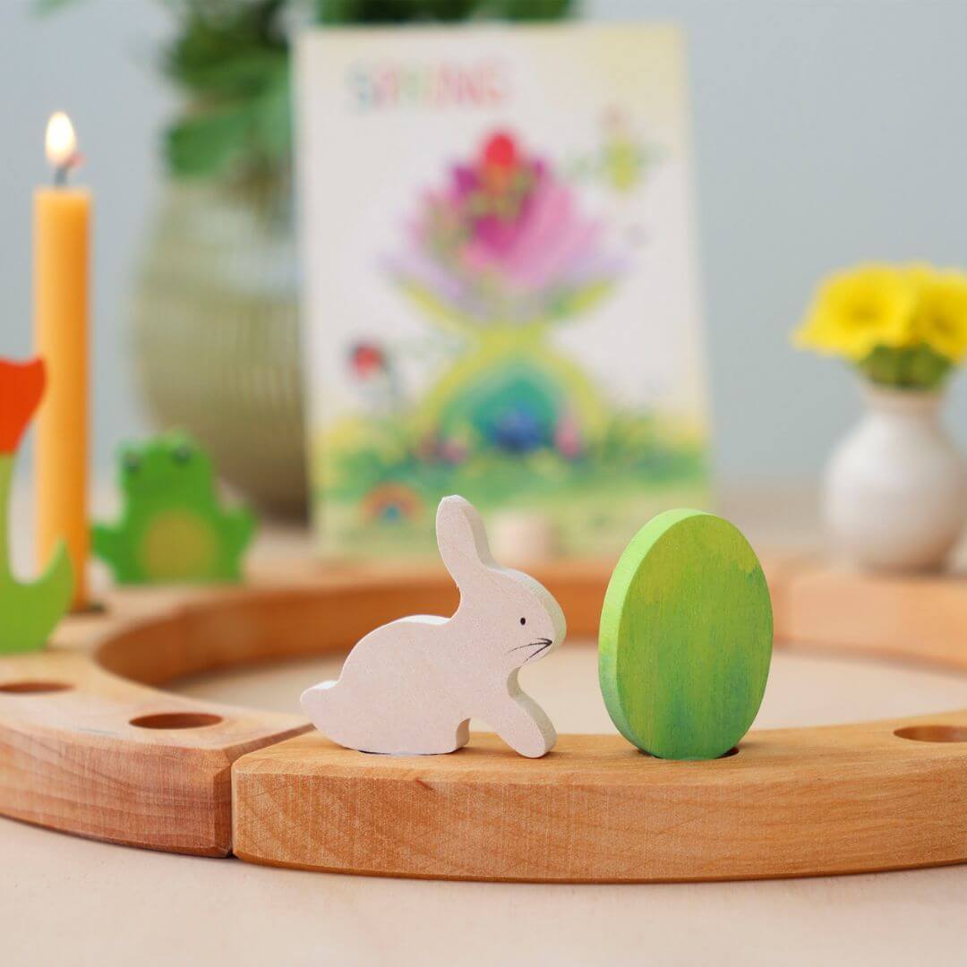 Grimm’s Wooden Easter Egg and Bunny in the green colorway displayed on a wooden celebration ring, accompanied by a yellow candle, a small vase with flowers, a wooden frog, a wooden red tulip, and festive decorations.