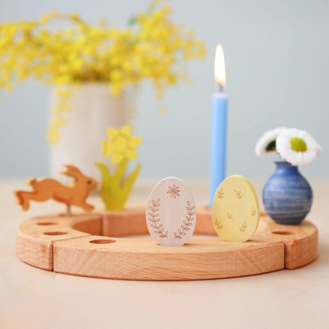 Grimm’s Wooden Easter Eggs Ornamental Colrways displayed on a wooden celebration ring, accompanied by a blue candle, a small vase with flowers, a wooden bunny, a wooden daffodil, and festive decorations.