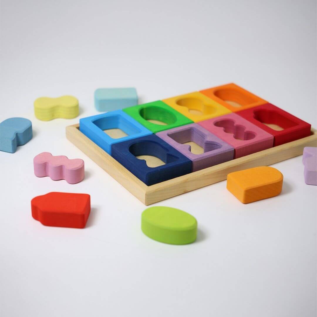 Grimm’s Windows Wooden Building Set displayed in a wooden tray with colorful shaped blocks in rainbow hues, some pieces scattered for open-ended play. Bella Luna Toys