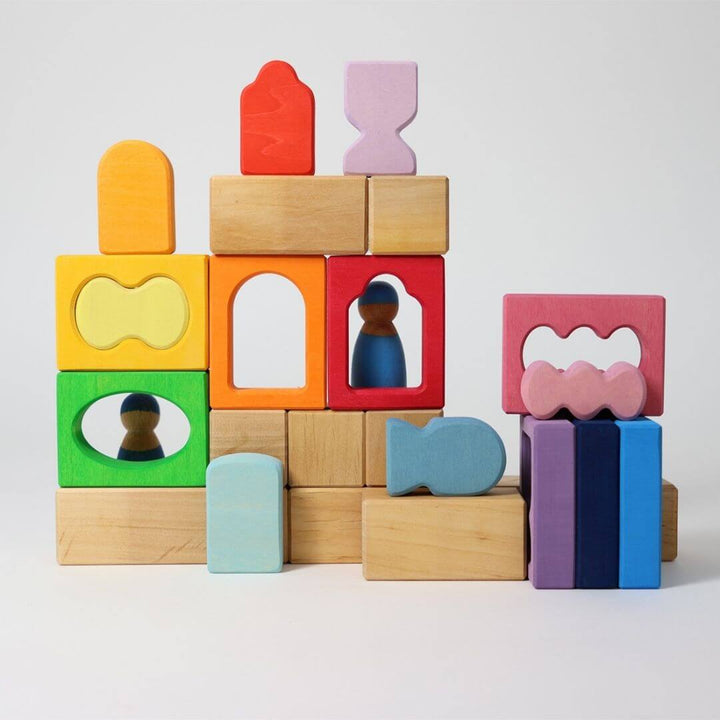 Grimm’s Windows Wooden Building Set stacked into a colorful block structure with natural wooden pieces and rainbow window shapes, featuring two small peg dolls inside for imaginative play. Bella Luna Toys