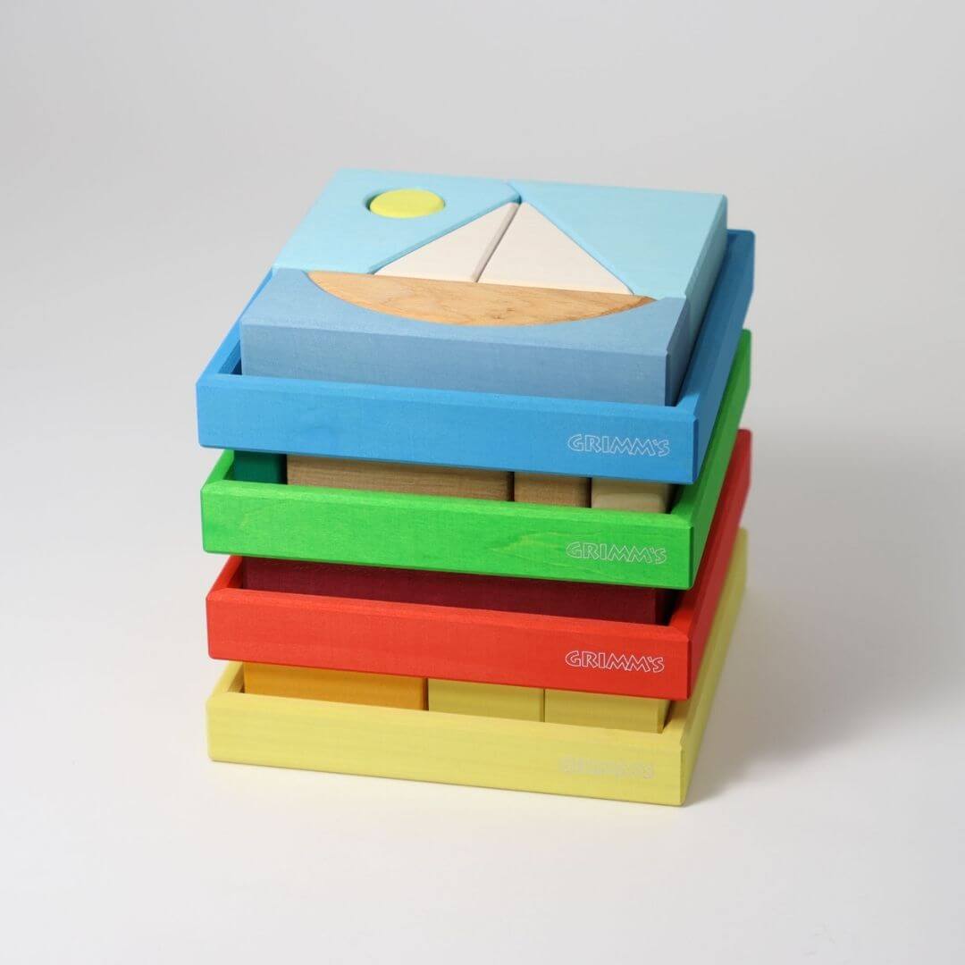 Four Grimm’s Wooden Puzzle and Building Sets in a wooden tray, stacked in a pile featuring colorful nature- and vehicle-inspired scenes in red, yellow, green, and blue for open-ended play. Bella Luna Toys