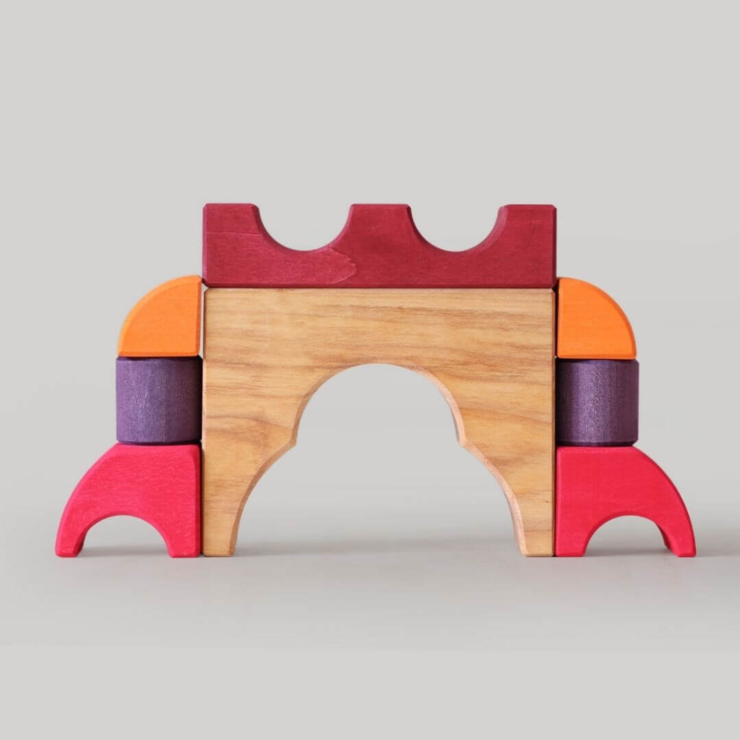 Grimm’s Wooden Puzzle and Building Set in red shown with organic-shaped pieces arranged outside the frame, forming playful sculptures and designs. Bella Luna Toys