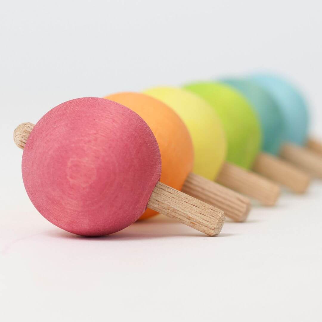 Side-view of six colorful wooden spinning tops arranged in a rainbow line, each with a round ball handle, crafted by Grimm’s.