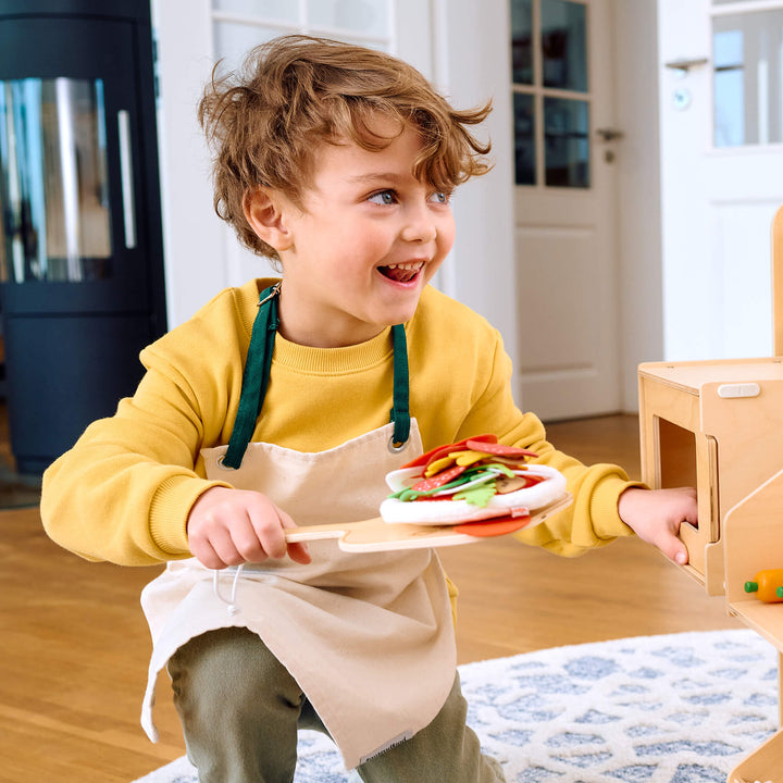 Child in a yellow shirt and apron holding a toy pizza by HABA in a home setting
