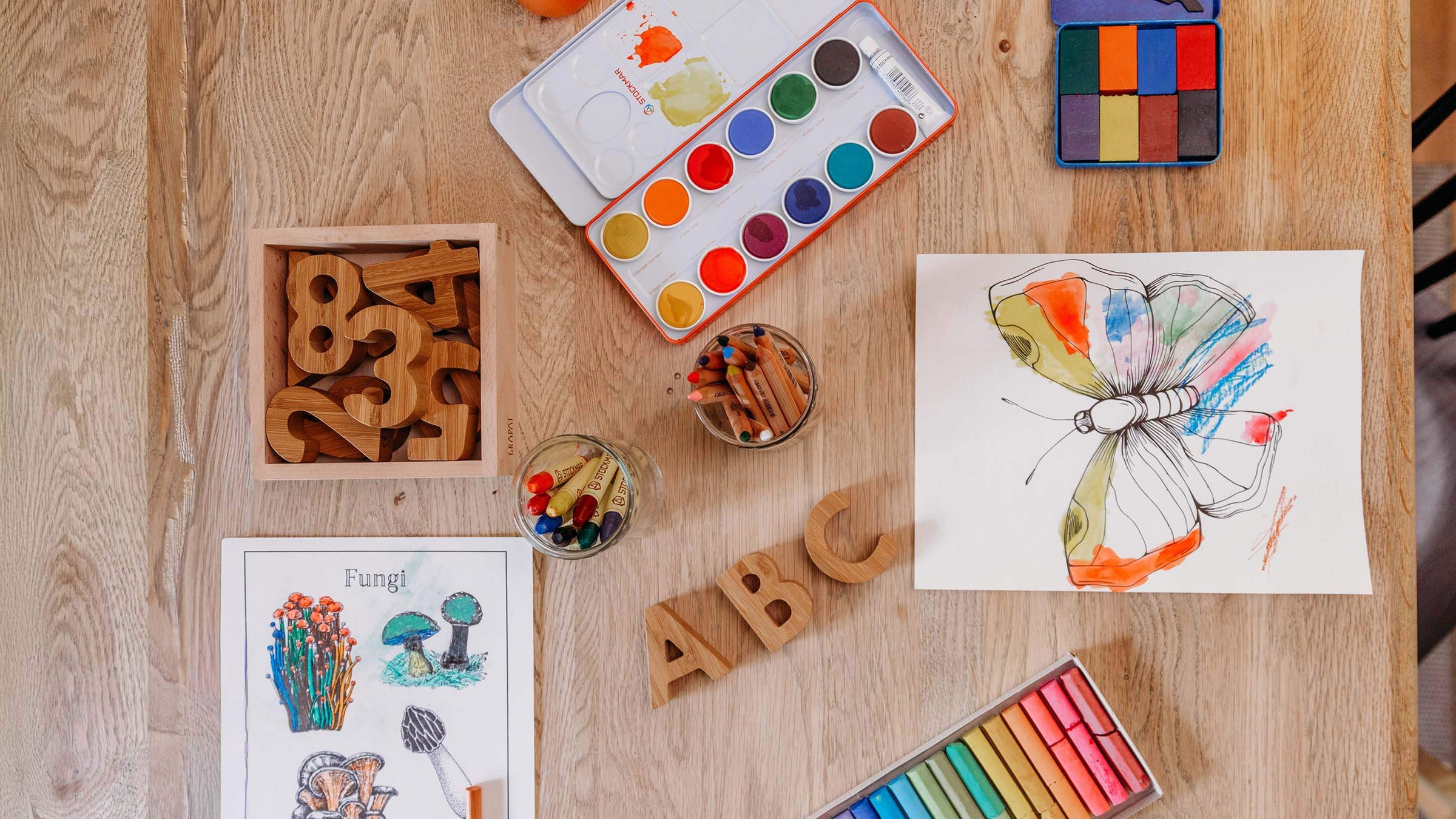 A styled flat-lay of children's art and learning supplies on a light wood table, including a butterfly watercolor painting, wooden number blocks, alphabet blocks, crayons, a watercolor paint palette, and an ABC chart.