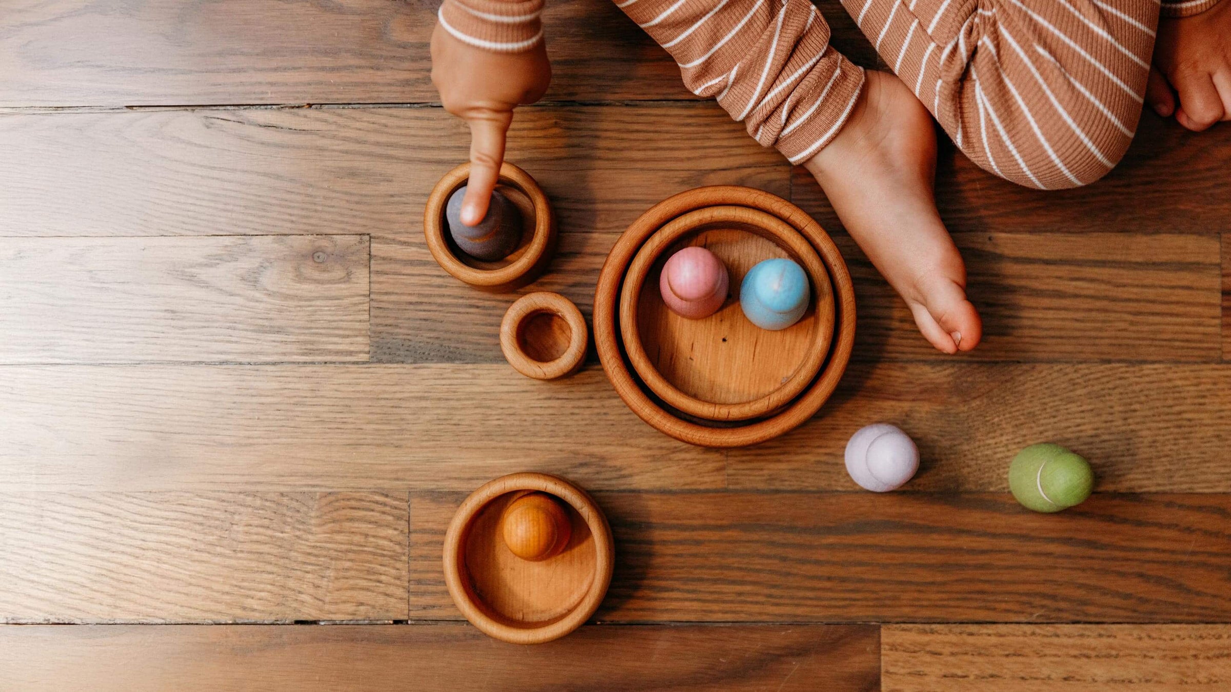 A child's hand and legs in striped pajamas interact with a set of wooden nesting bowls and colorful painted balls on a wooden floor.