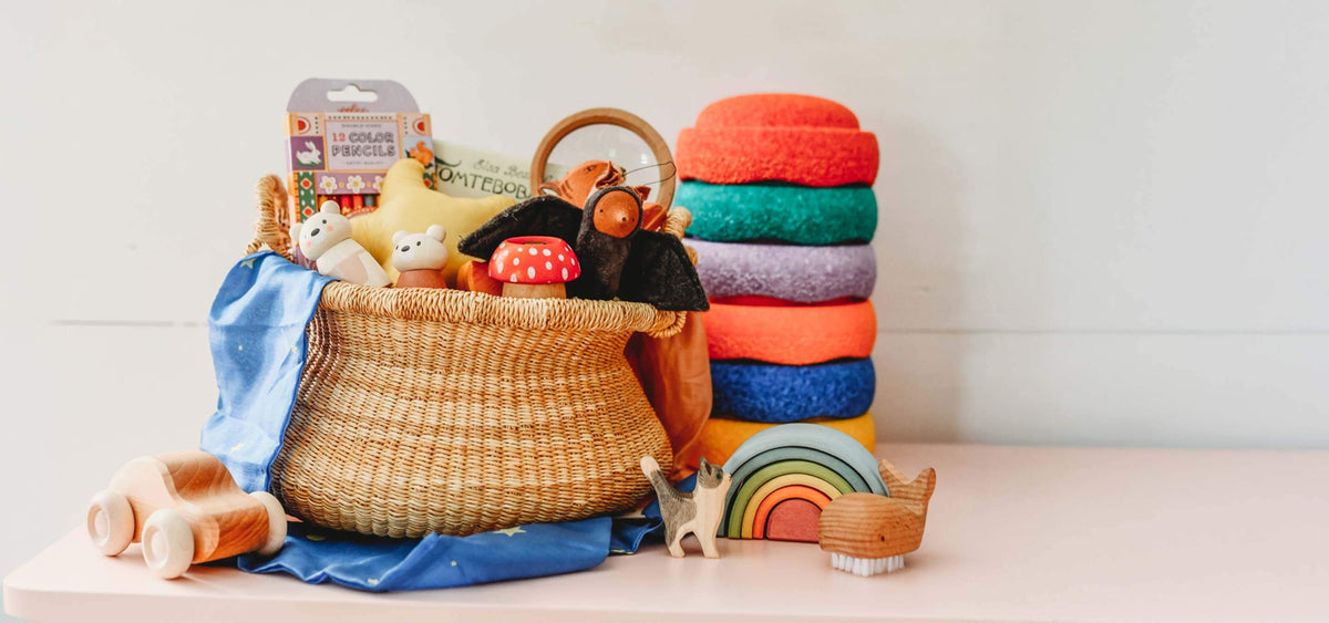 A styled collection of children's toys and accessories arranged on a light pink surface against a white wall. The set includes a woven basket filled with plush toys and a packaged item, a stack of colorful felt washcloths, wooden rolling pins, a wooden rainbow toy, and a small wooden animal figure.