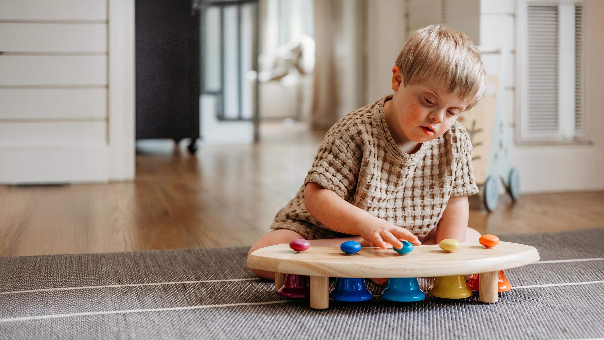 A young child sits on a woven rug in a bright room, playing with a small wooden toy table featuring colorful bells and pegs.