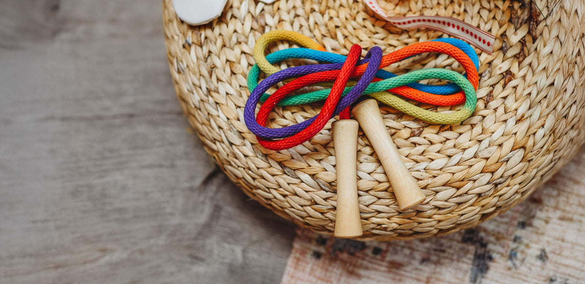 A colorful jump rope with wooden handles resting on a woven straw hat, set against a rustic gray wooden background.