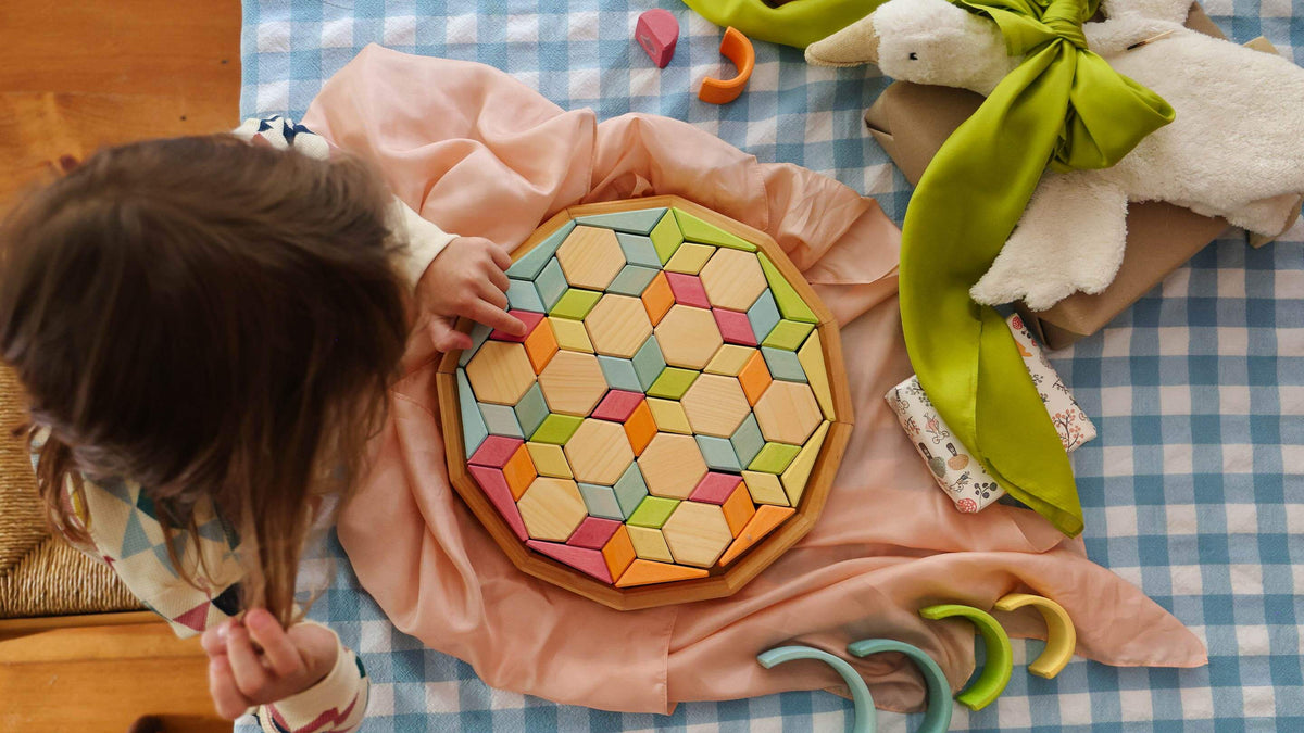 A child's hand touches a colorful geometric wooden puzzle board on a pink cloth, set on a blue-and-white check blanket with toys nearby.