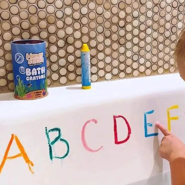 Child writing letters with bath crayons on a bathtub, with Honeysticks beeswax bath crayons visible.