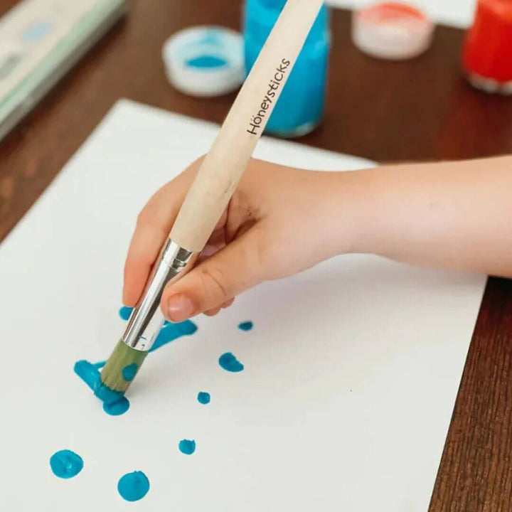 Hand holding a paintbrush with 'Honeysticks' branding, painting blue dots on white paper.