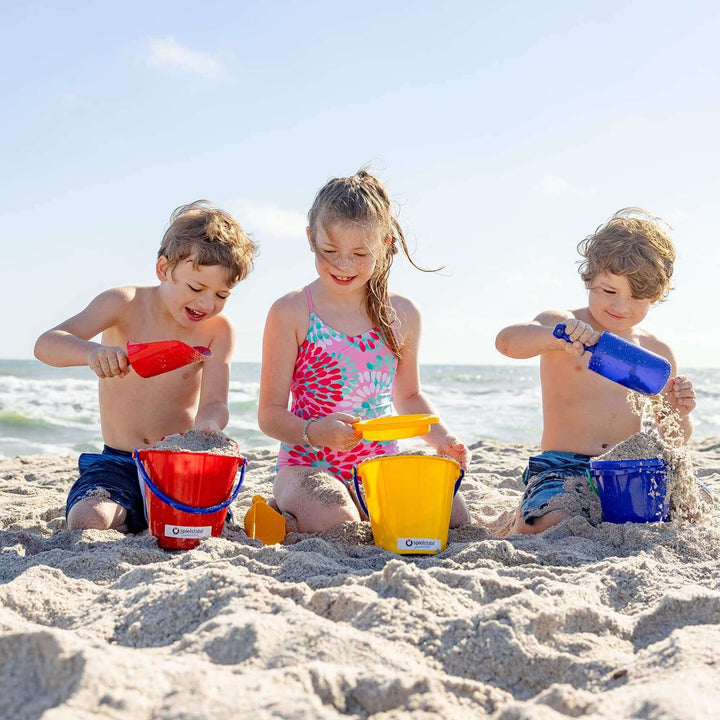 3 children kneeling on the beach playing with red, yellow, and blue Spielstabil buckets, red and blue sand scoops, and a yellow sand sieve